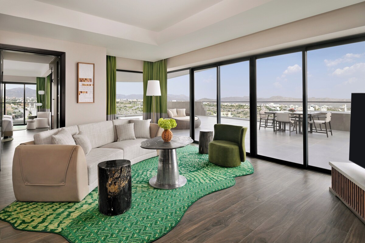 Un salon moderne avec un canapé, des fauteuils, une table basse et une vue sur la ville grâce à de grandes fenêtres.