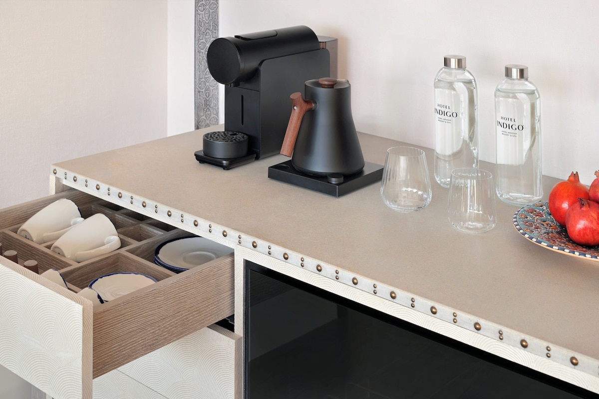 Una encimera de cocina con una cafetera, dos botellas de vidrio con la etiqueta "Hotel Indigo", un plato con higos chumbos y un cajón con tazas y un hervidor.