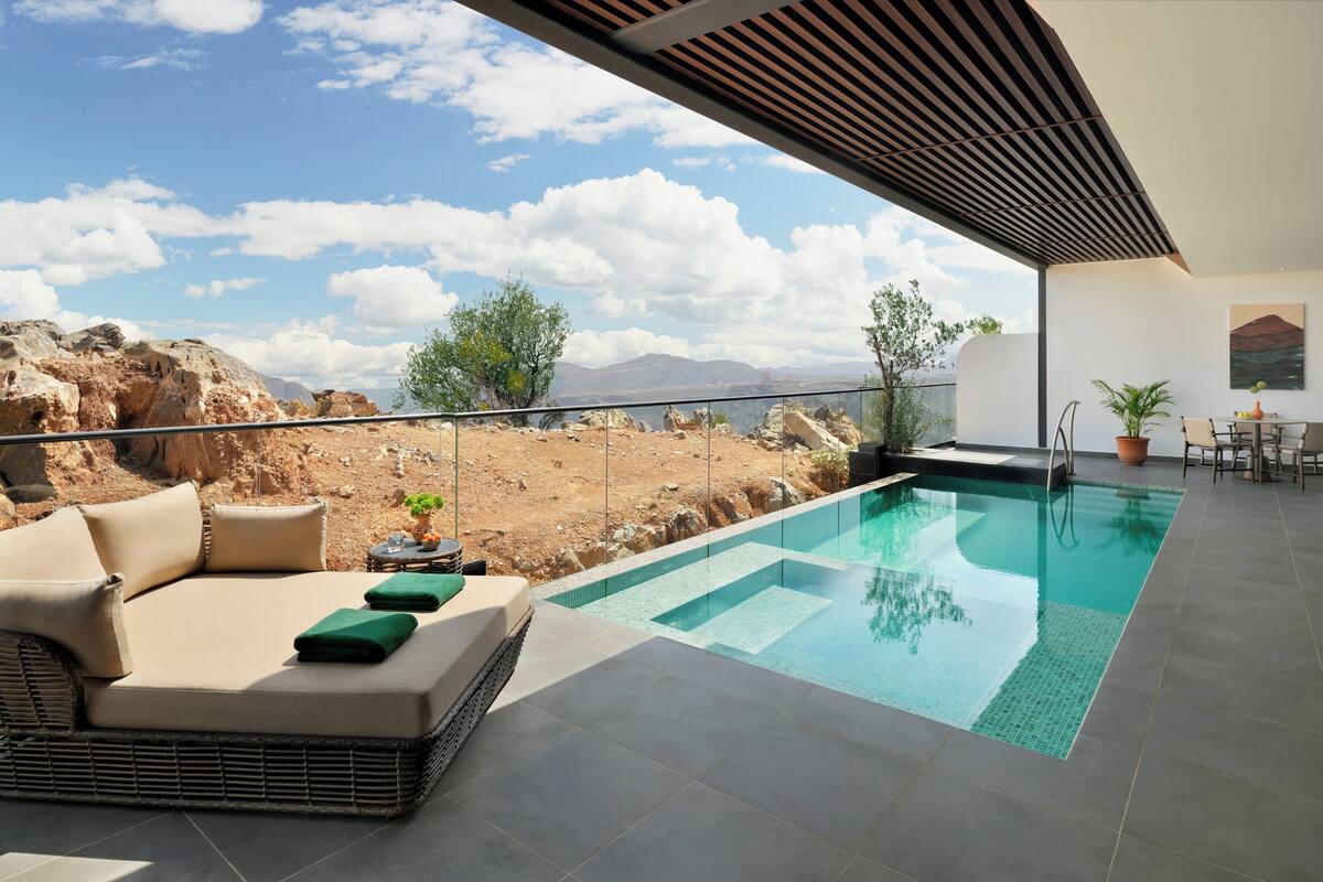 Un espace piscine extérieur moderne, doté d’une chaise longue et de coussins, offrant une vue sur le paysage désertique, ainsi qu’un coin repas avec une table et des chaises.