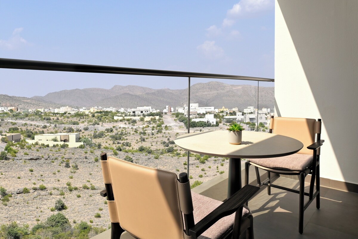 Un balcon avec une table et deux chaises, donnant sur un paysage désertique avec des bâtiments visibles au loin.