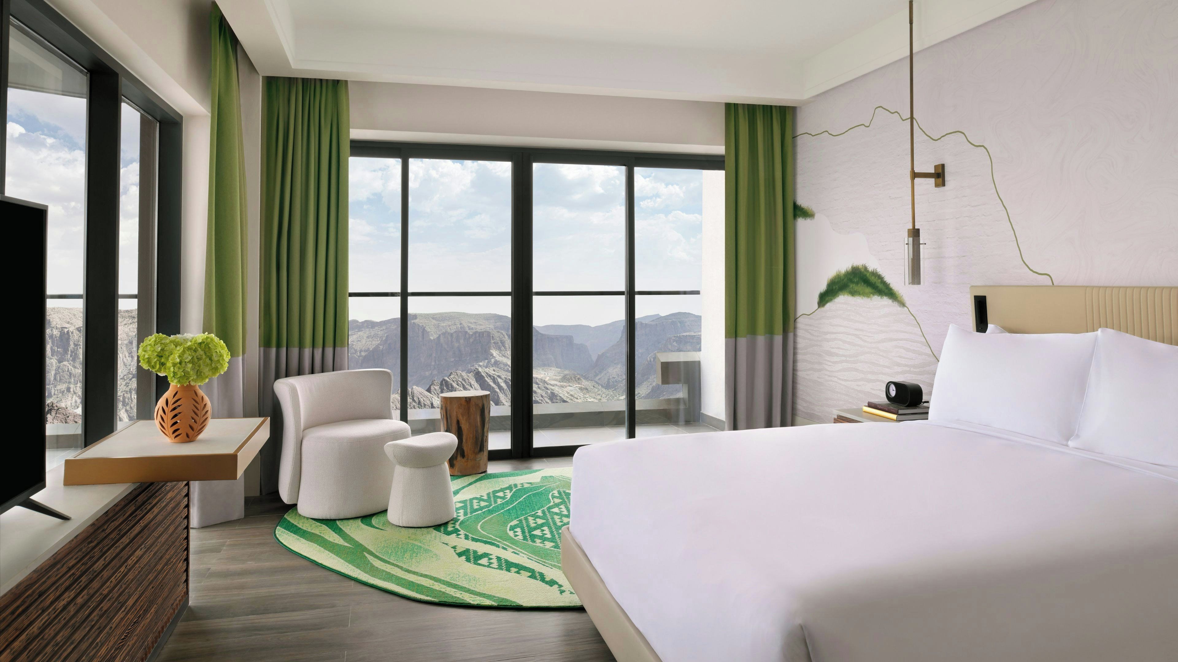 A hotel room with a large bed, a chair, a small table, a TV, and a view of a mountainous landscape through large windows.