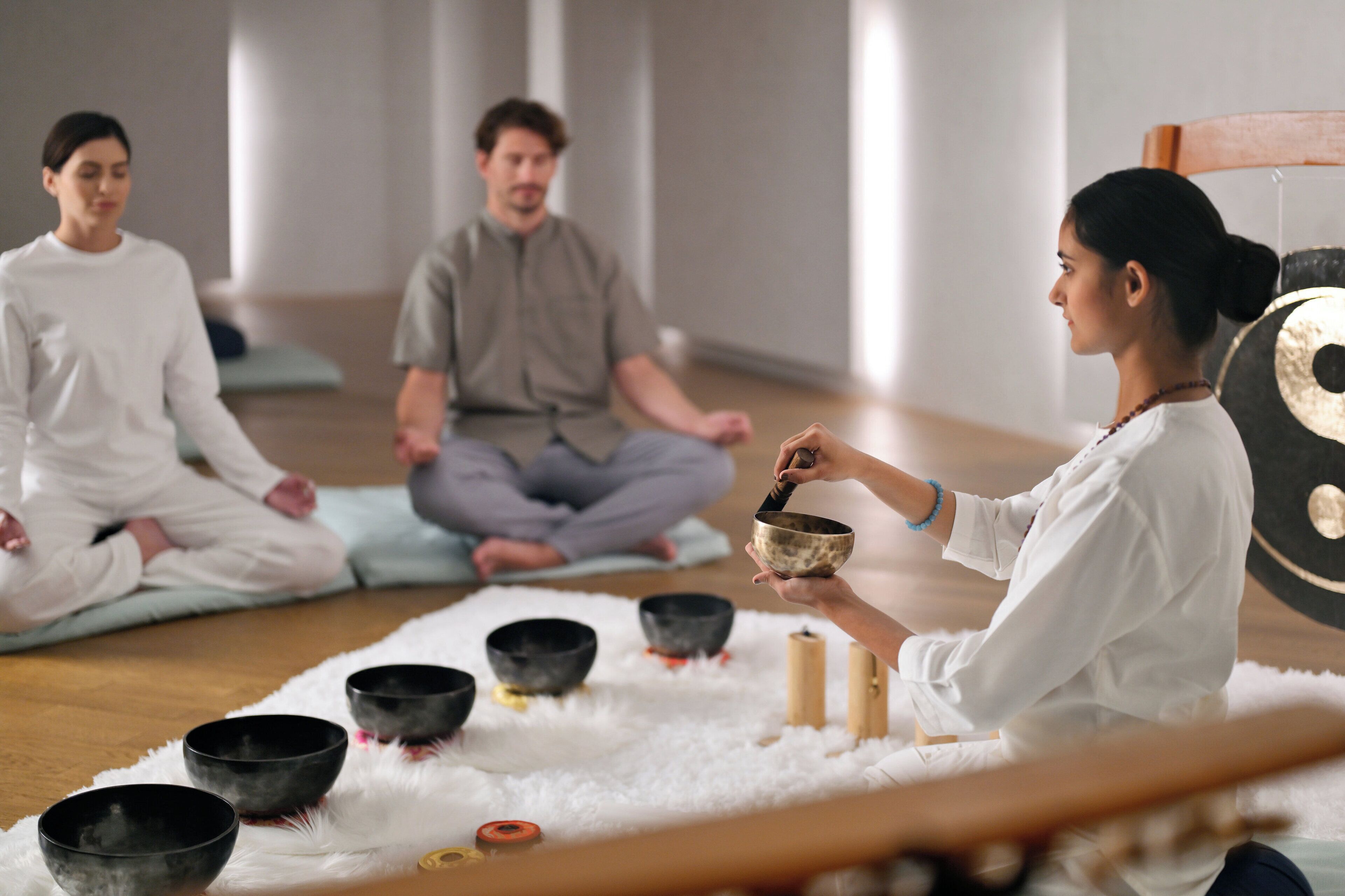 Een groep mensen mediteert in een ruimte met een zachte, witte vloerkleden, zwarte meditatiekommen en een houten stoel.