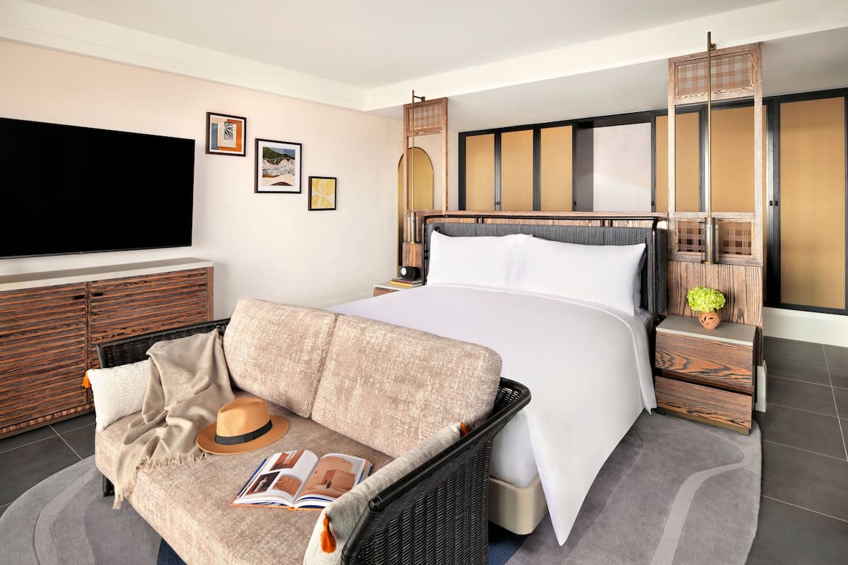 Une chambre d’hôtel moderne dotée d’un grand lit, d’une télévision à écran plat, d’un canapé et d’une petite table sur laquelle se trouvent un chapeau et un magazine.