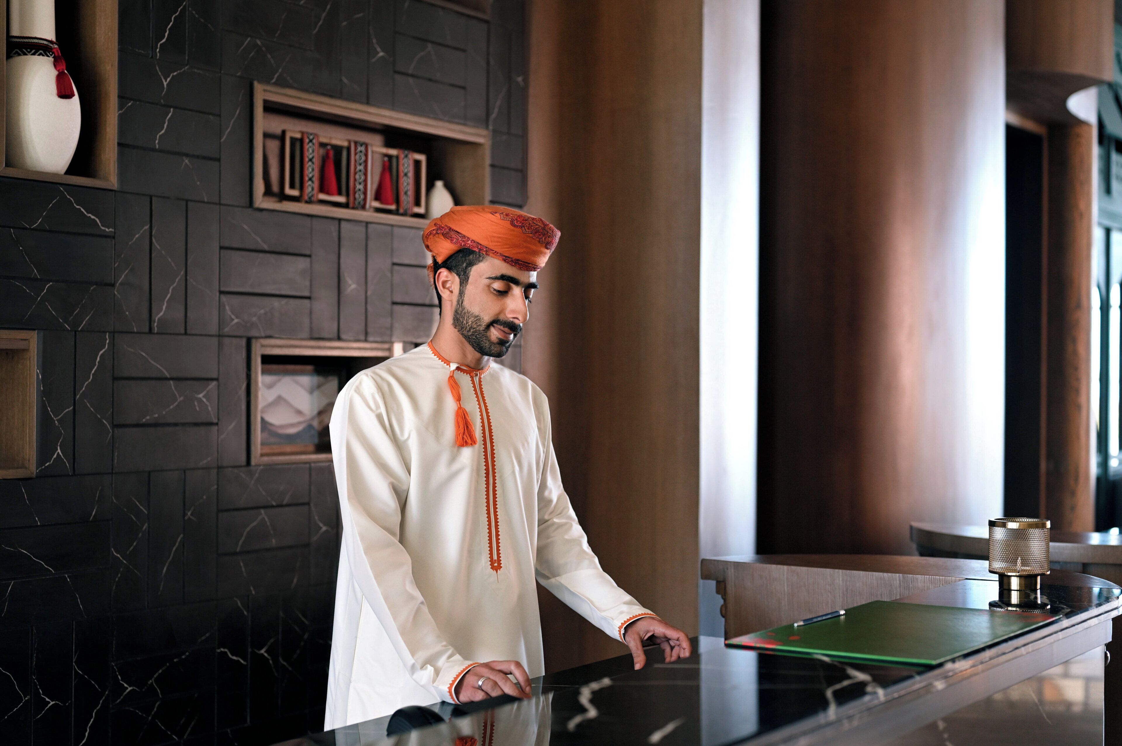 A man in traditional attire standing by a counter with a modern interior design, including marble walls and wooden accents.