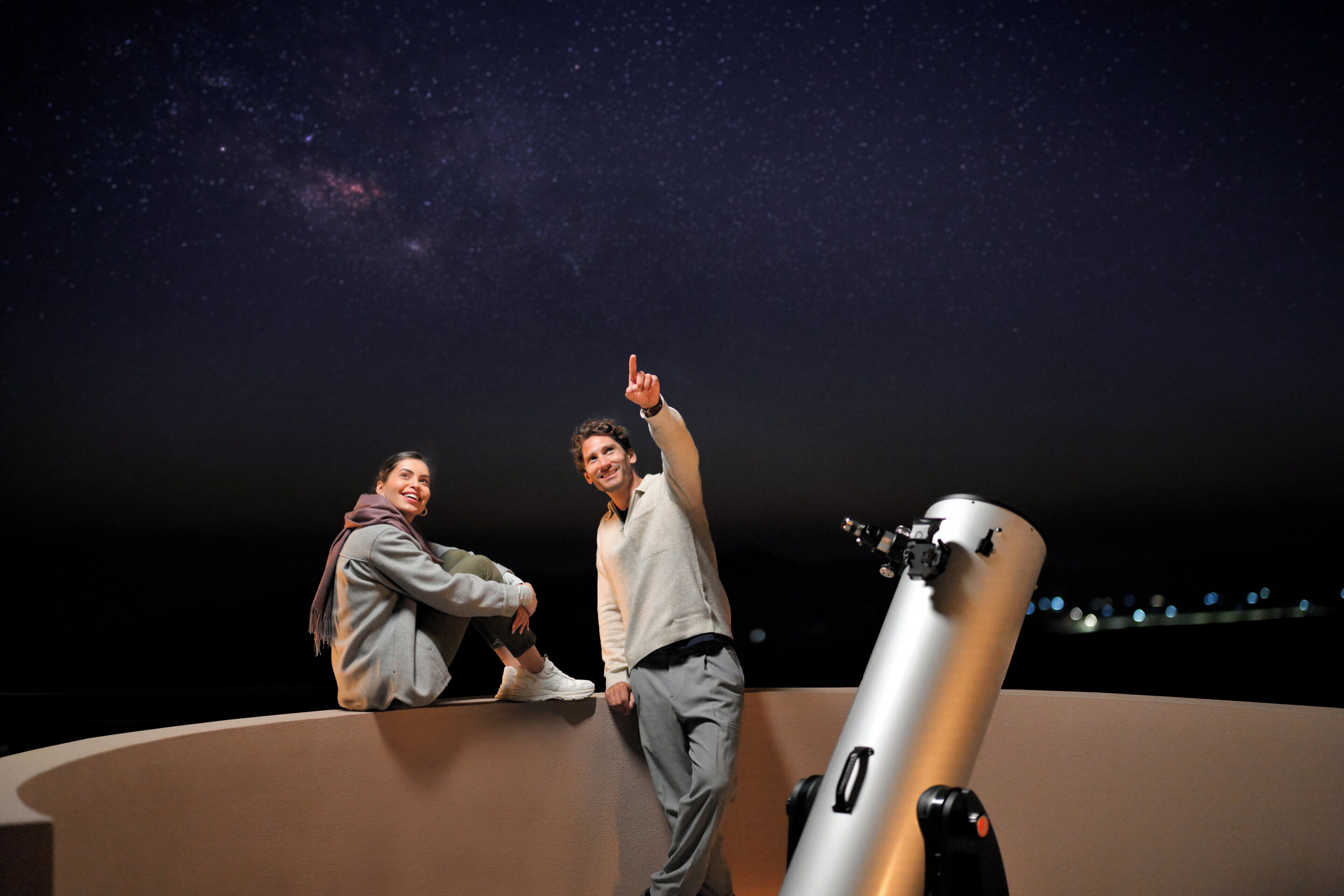 Deux personnes se tiennent sur une plateforme incurvée, un télescope à proximité, et un ciel étoilé en arrière-plan.