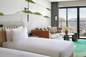 Habitación de hotel con una cama grande, un sofá, una mesita redonda y vistas a las montañas a través de amplias ventanas.