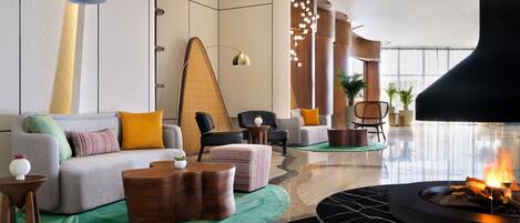 A modern lounge area with a sofa, ottoman, side tables, a fireplace, and a large window.