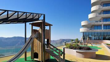 Un parque infantil con tobogán y estructura de escalada, un edificio con varios balcones y vistas a montañas distantes.