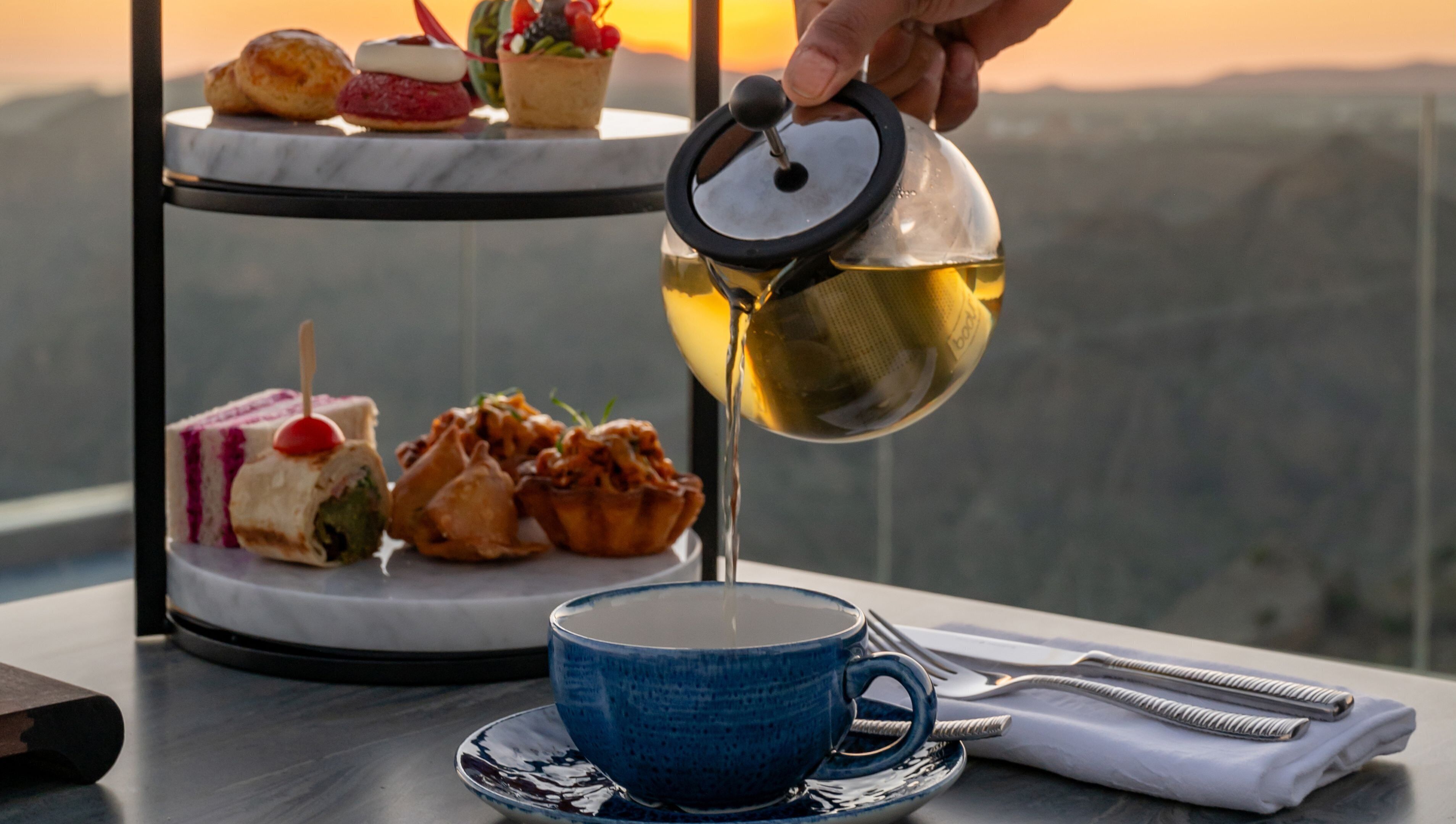 A tea set with a cup and saucer, a teapot, and a tray with pastries and sandwiches.
