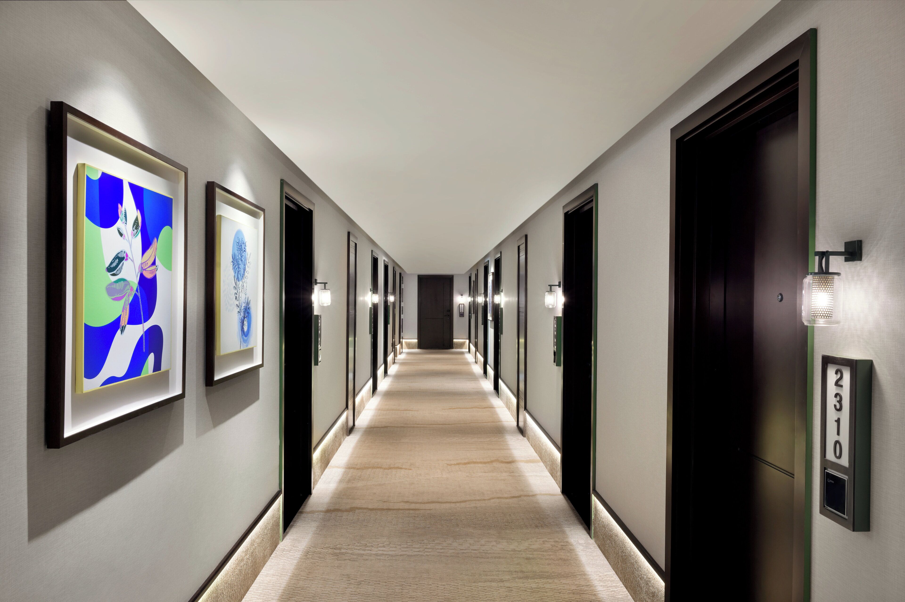 A hotel hallway with framed artwork on the walls, a carpeted floor, and doors with numberplates.