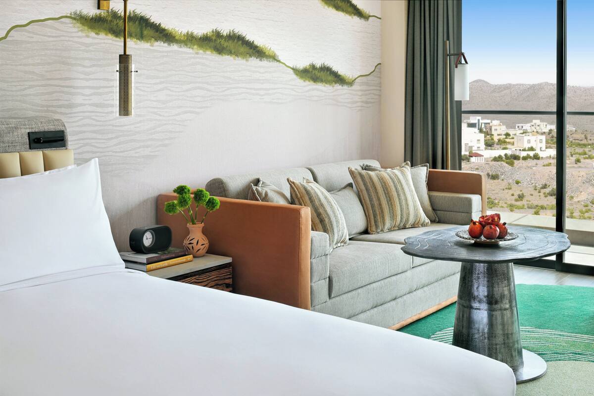 Une chambre d’hôtel moderne avec un lit, un canapé, une table ronde agrémentée d’une plante en pot et une vue sur le paysage désertique.