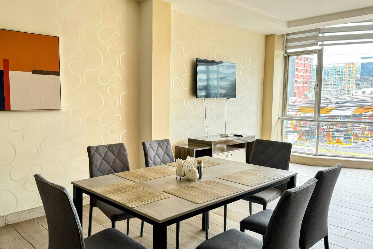 A modern hotel room with a dining area, a flat-screen TV mounted on the wall, and a large window offering a city view.