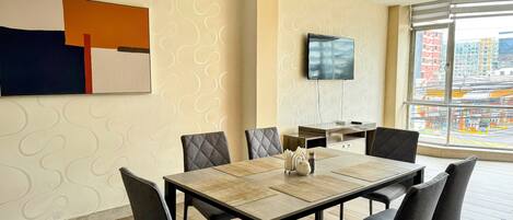 A modern hotel room with a dining area, a flat-screen TV mounted on the wall, and a large window offering a city view.