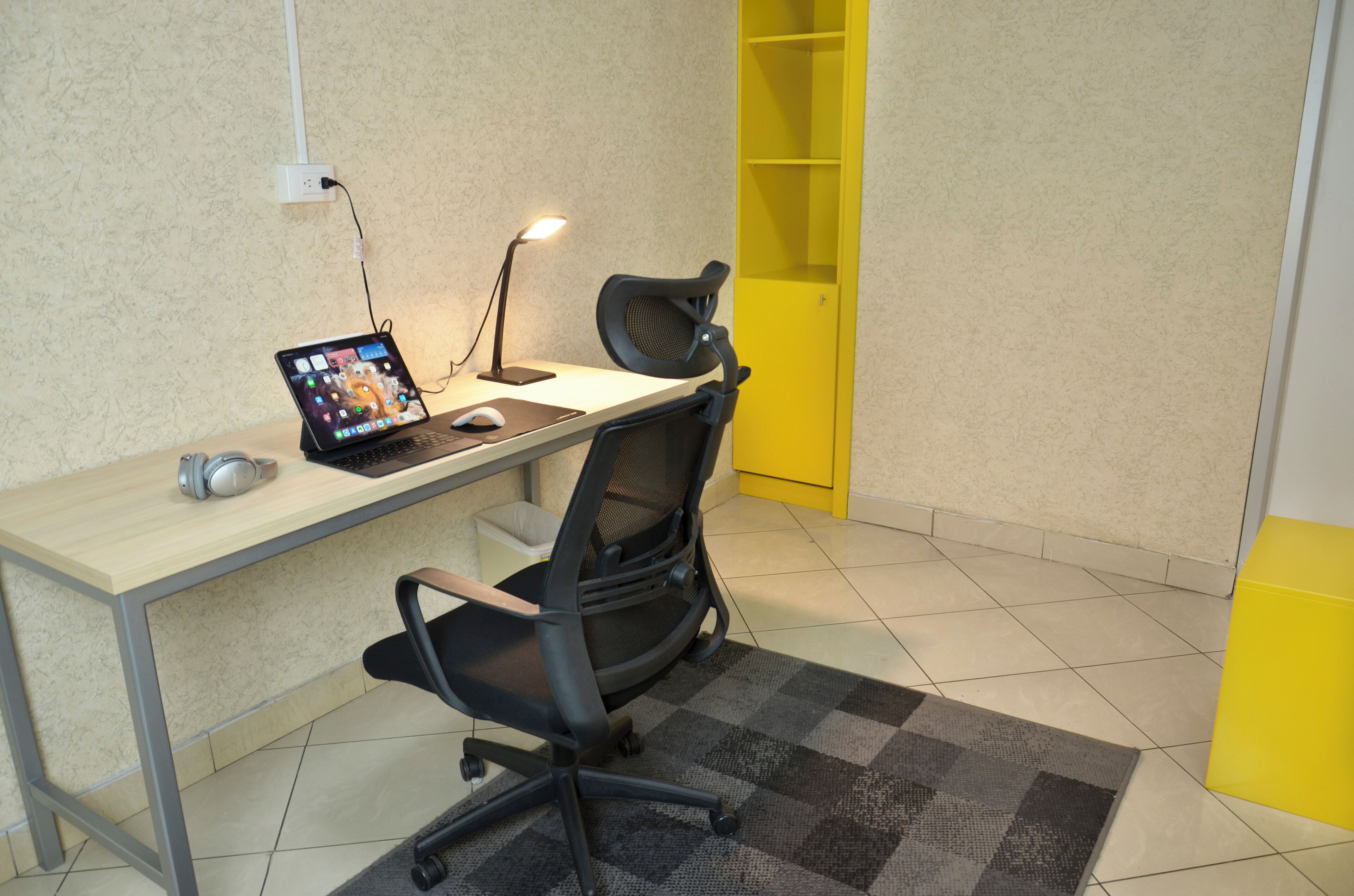 A workspace with a desk, two chairs, a laptop, a mouse, a lamp, and a yellow storage unit.