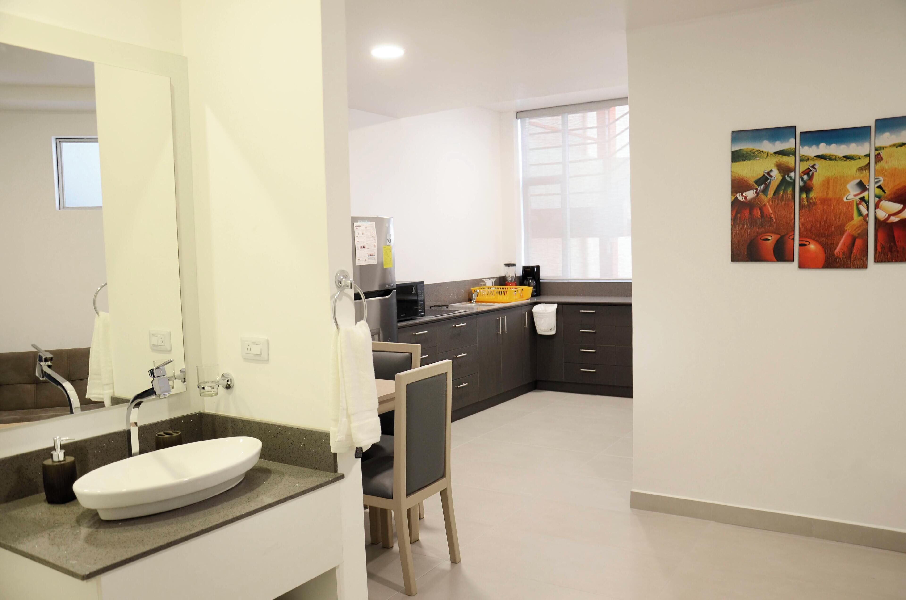 A modern bathroom with a large mirror, a white sink, and a dark countertop. There is a chair and a small table with a towel.