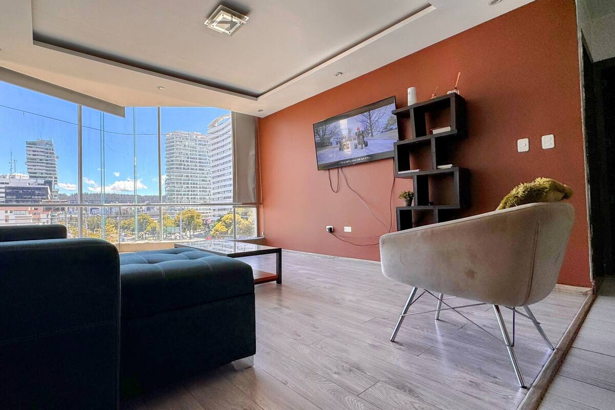 A modern living room with a large window, a flat-screen TV mounted on the wall, and a minimalist furniture arrangement.