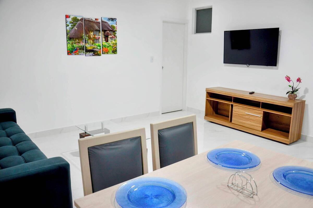 A modern living room with a sofa, a TV mounted on the wall, a wooden cabinet, and a dining table with blue plates.