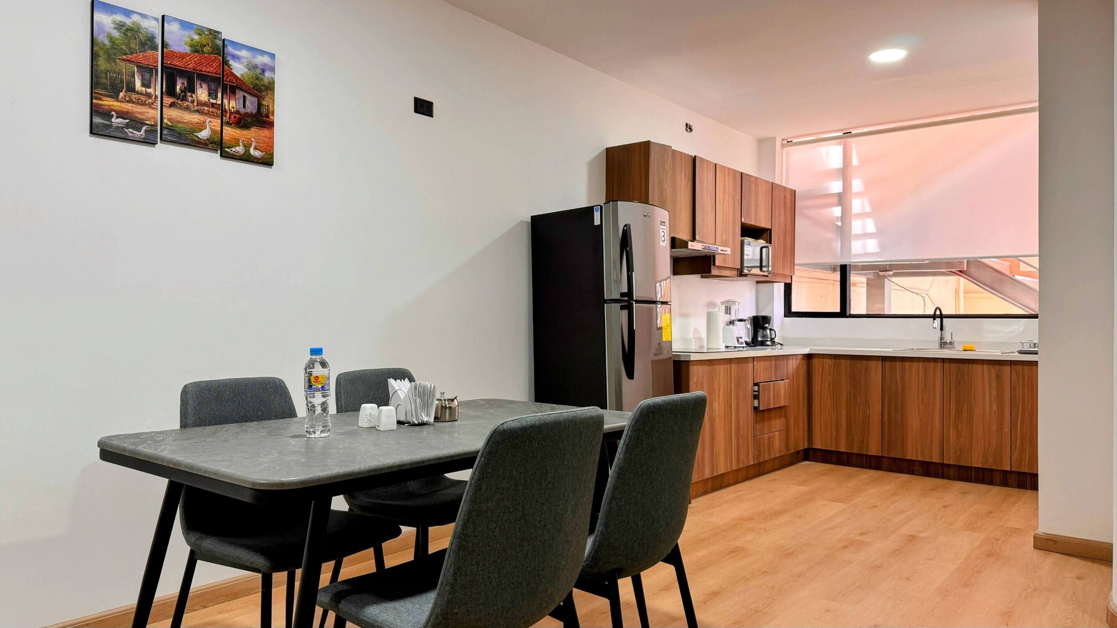 A compact kitchen with wooden cabinets, a refrigerator, and a sink. There is a dining table with chairs and a water bottle on it.