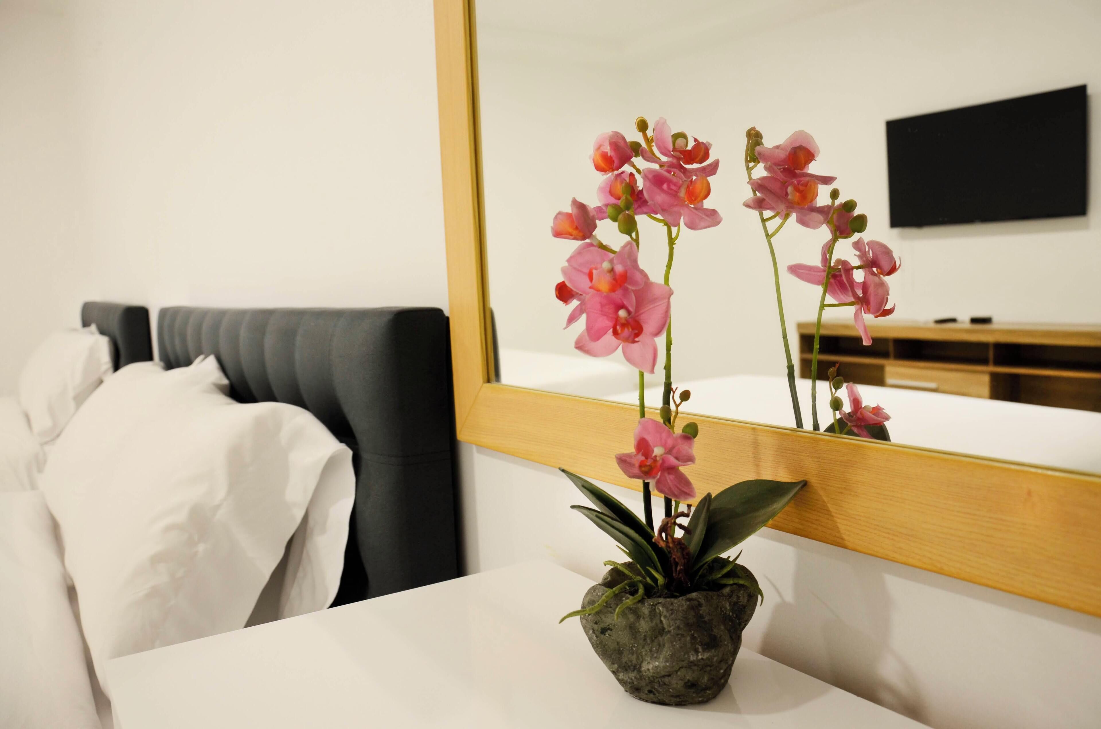 Habitación de hotel con cama, mesita de noche blanca con orquídea en maceta y un espejo grande que refleja la habitación.