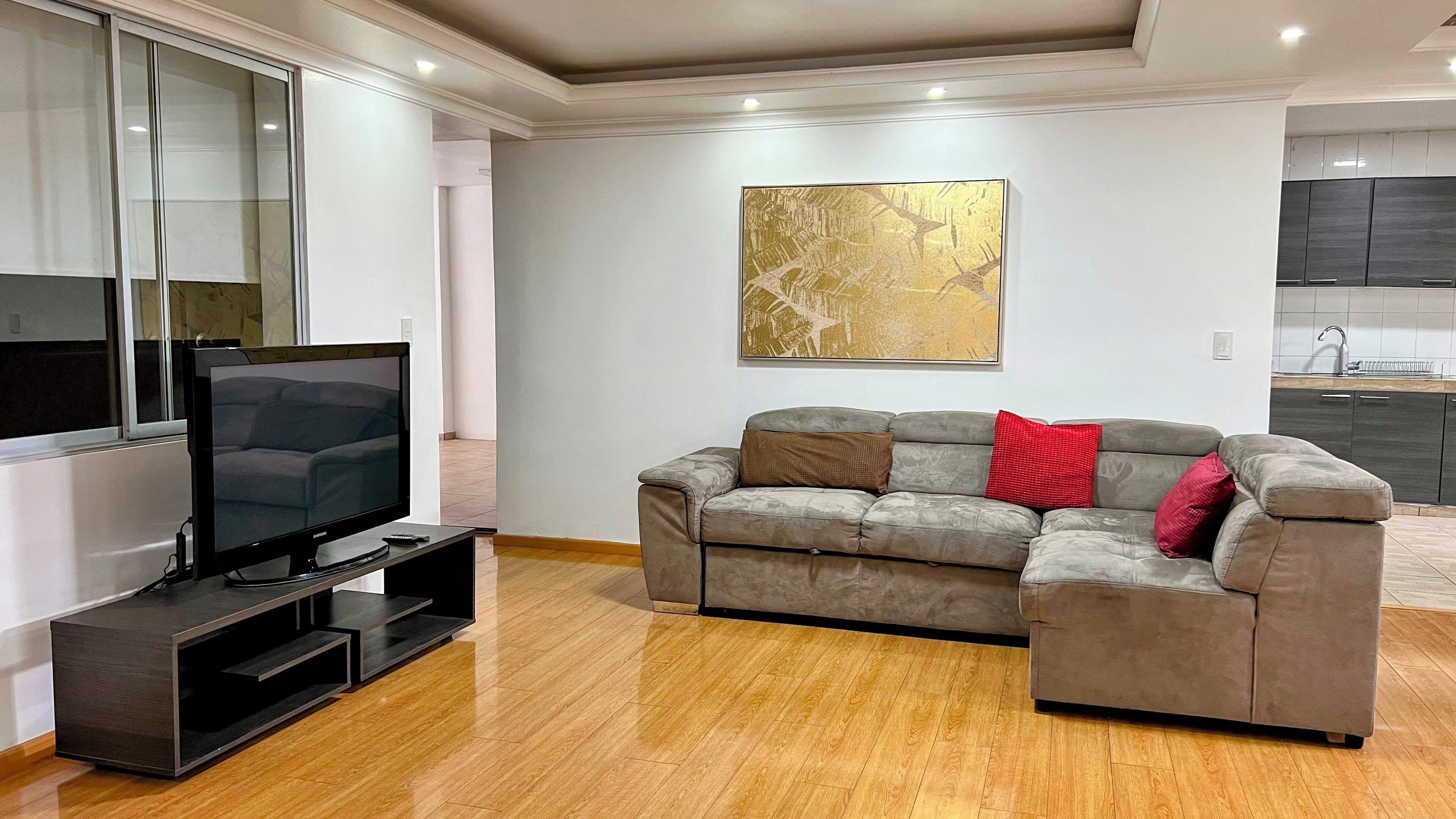 A modern living room with a flat-screen TV on a stand, a grey sectional sofa with red and brown cushions, a large abstract gold painting on the wall, and a kitchen area in the background.