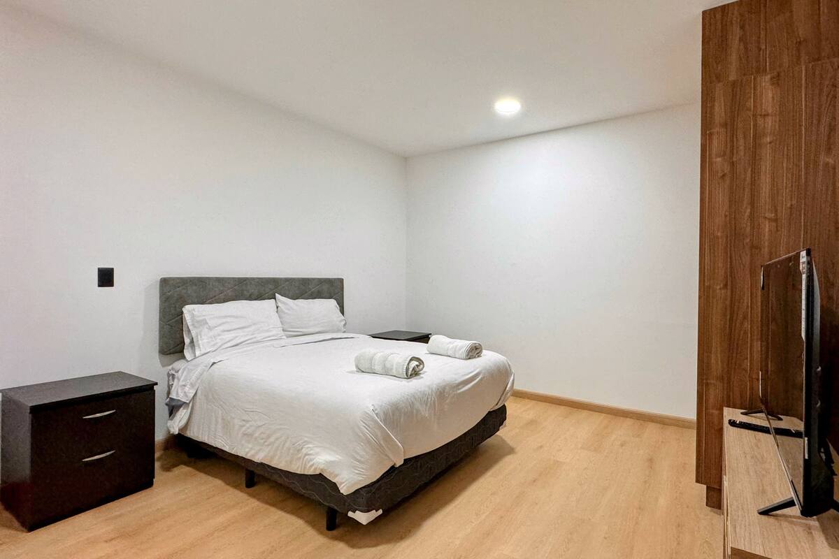 A bedroom with a bed, a nightstand, a wooden wardrobe, and a TV.