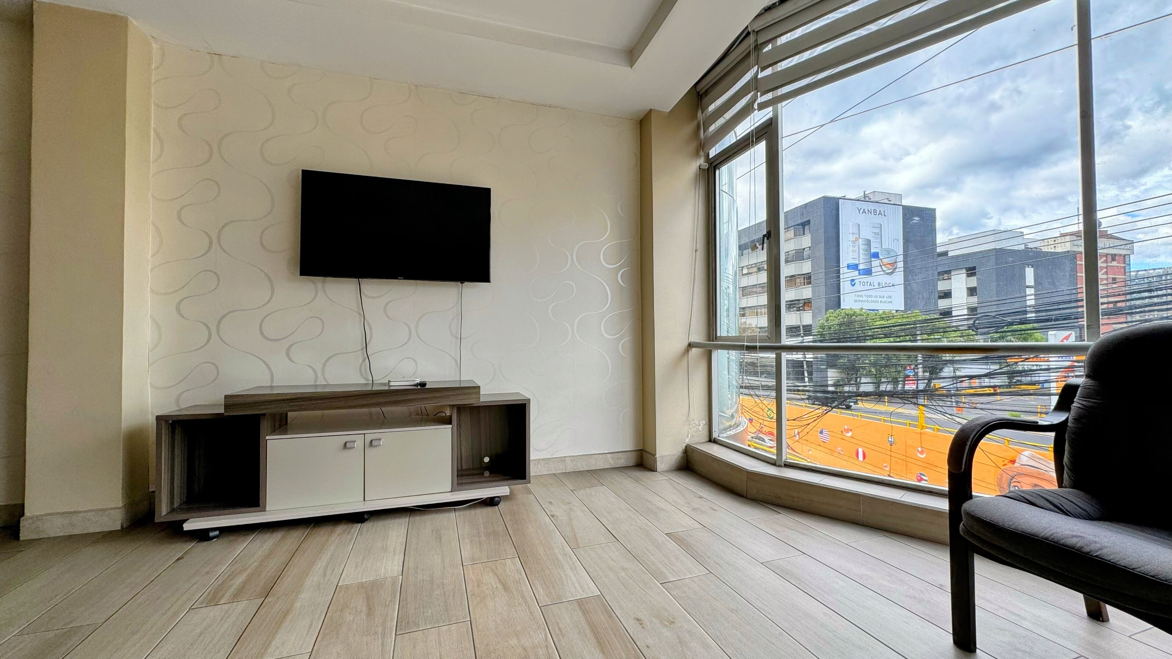 A modern living room with a flat-screen TV mounted on the wall, a wooden entertainment unit, and a large window offering a view of a cityscape.