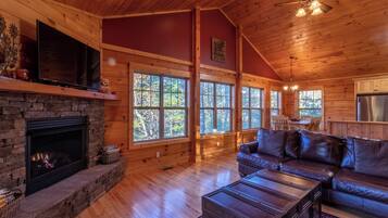 A spacious living room with a stone fireplace, leather sofa, and large windows offering a view of the forest.