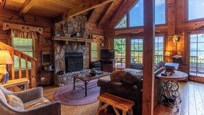A log cabin interior with a stone fireplace, wooden furniture, and large windows offering scenic views. - Apple of my Eye (Banner Elk)