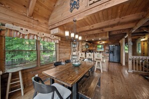 A wooden interior with a dining table, chairs, a kitchen area, and a large window with blinds. - Apple of my Eye (Banner Elk)