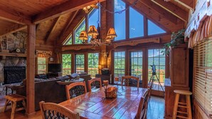 A rustic cabin interior with a wooden dining table, chairs, a fireplace, and large windows offering a view of the outdoors. - Apple of my Eye (Banner Elk)