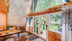 Een gezellig interieur van een vakantiehuisje met een houten plafond, een bank met gestreepte kussens, een bijzettafeltje en een groot raam met uitzicht op groen.