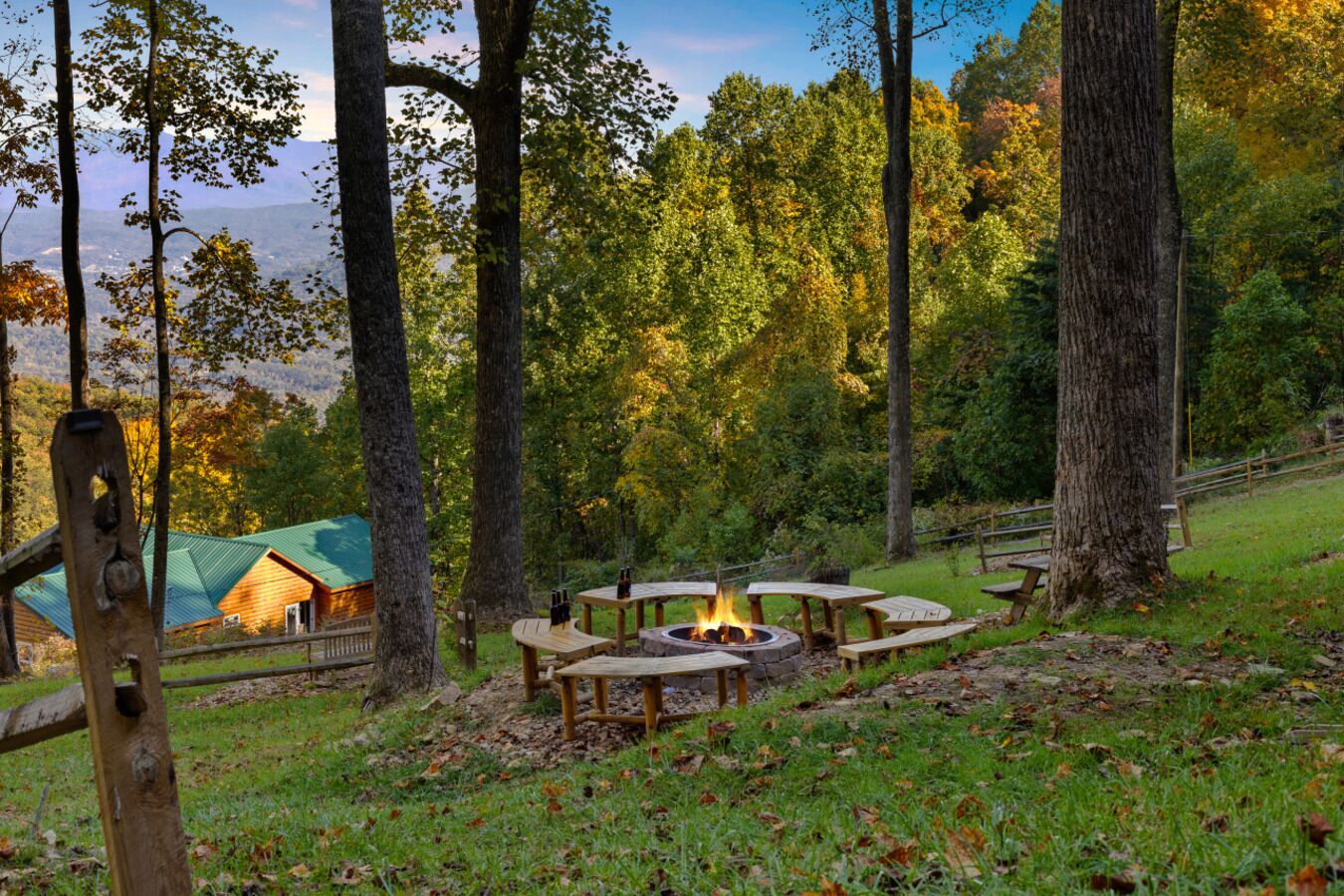 A campsite with a fire pit surrounded by picnic tables, a wooden cabin, and a forested backdrop.