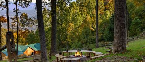 A campsite with a fire pit surrounded by picnic tables, a wooden cabin, and a forested backdrop.