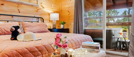 A cozy wooden cabin bedroom with a bed, a nightstand, a lamp, a teddy bear, a hat, a tray with snacks, and a book titled "Delicious Places".