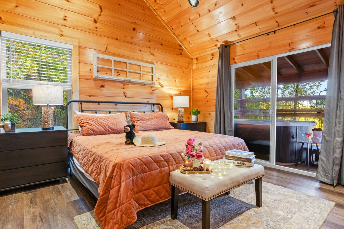 A cozy wooden cabin bedroom with a bed, bedside lamps, a small table with a hat and flowers, and a view of the outdoors.