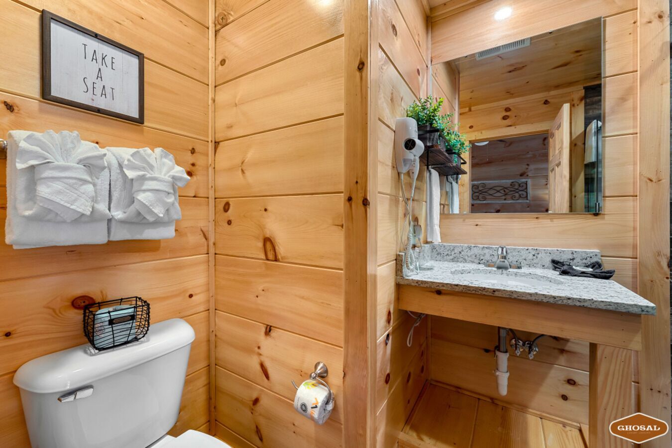 A wooden cabin-style bathroom with a toilet, a sink with a mirror, and a sign that says "TAKE A SEAT".