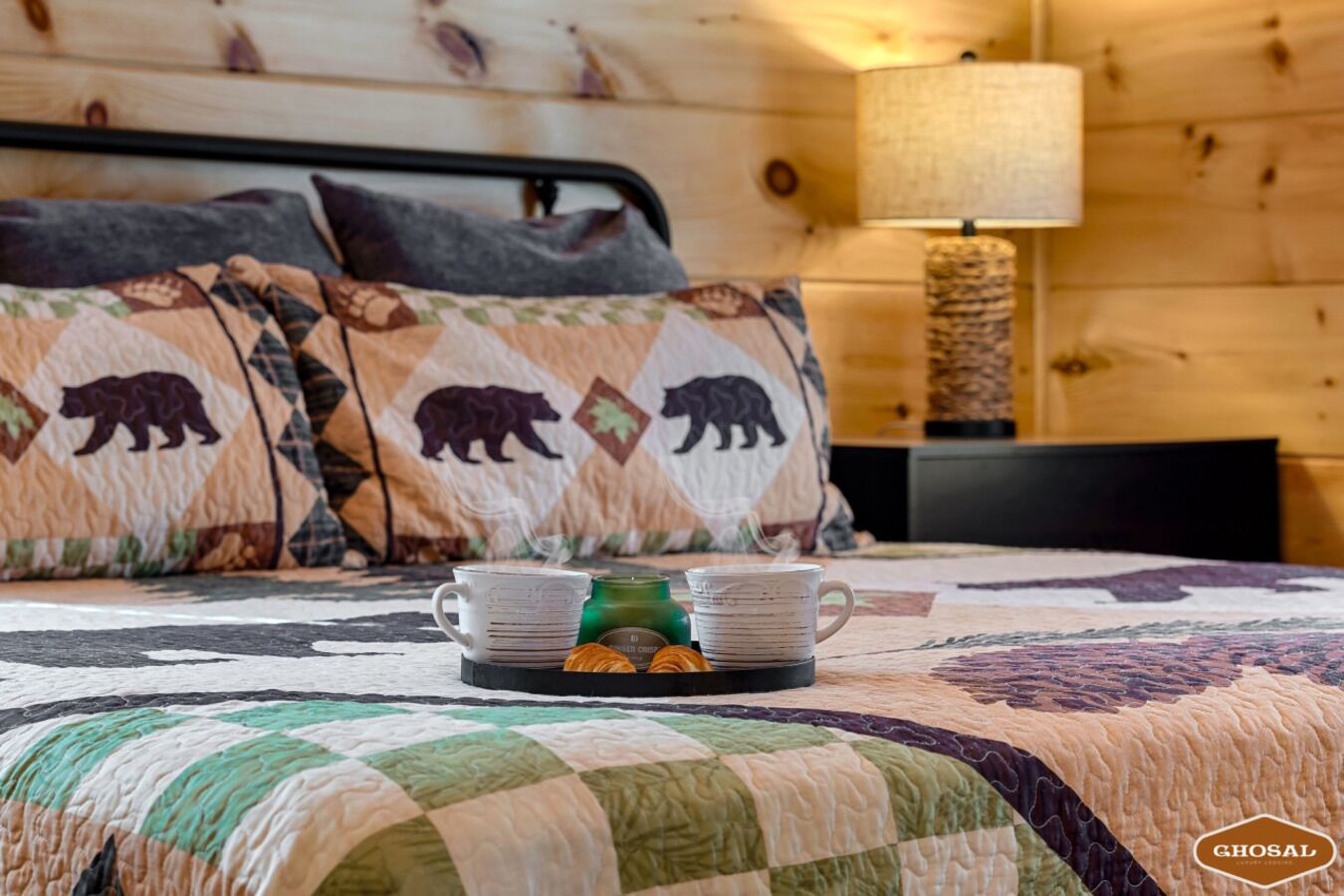 Um quarto aconchegante com colcha acolchoada, duas almofadas com estampas de ursos, uma luminária de cabeceira e um tabuleiro com duas canecas e uma vela.