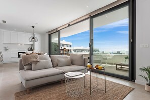 Ein modernes Wohnzimmer mit einer Couch, einem Couchtisch und einem Laptop. Ein großes Fenster bietet Blick auf Gebäude und einen klaren Himmel.