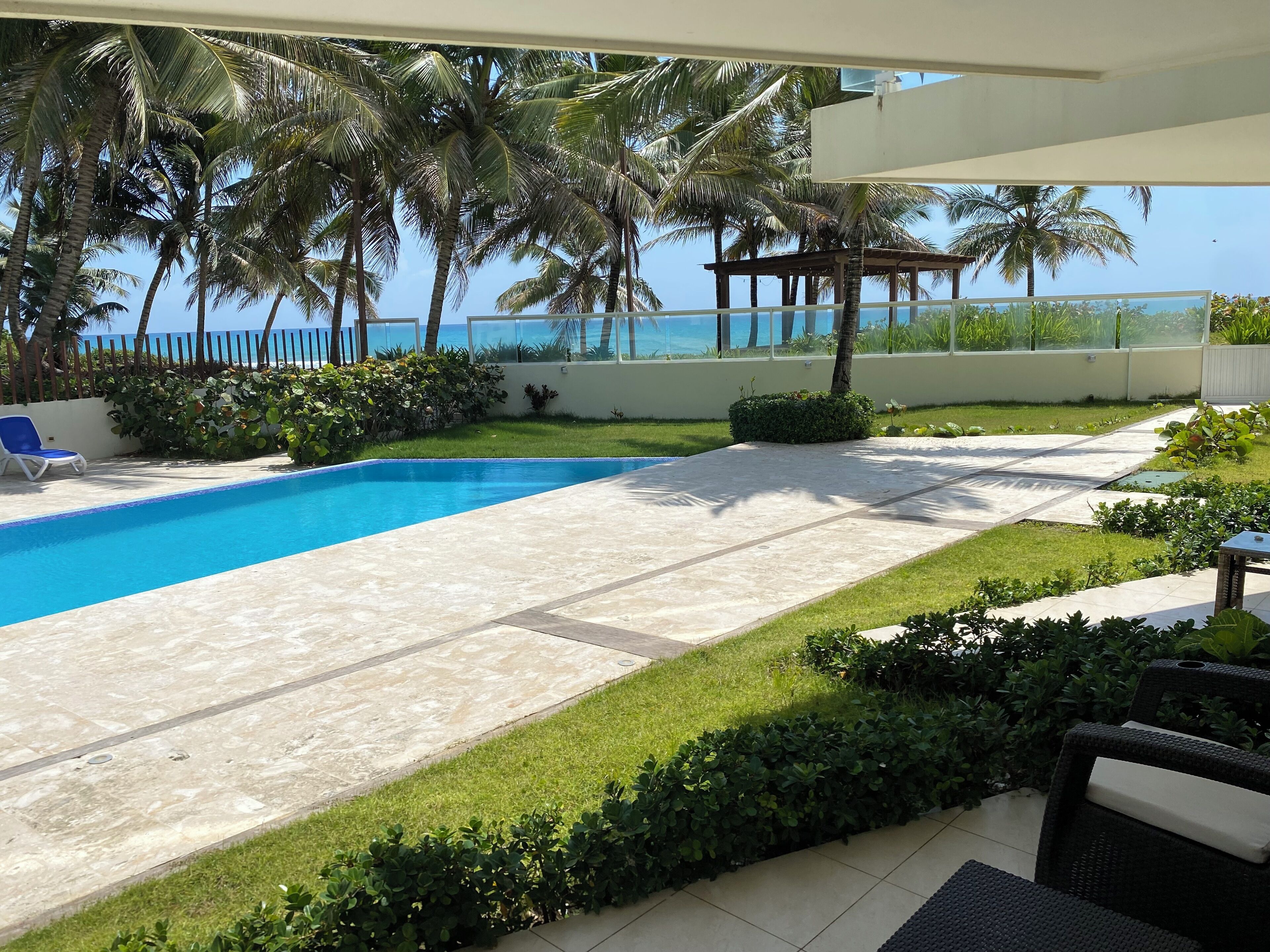 A pool area with a view of the ocean, surrounded by palm trees and a covered seating area.