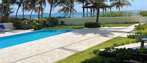 A pool area with a view of the ocean, surrounded by palm trees and a covered seating area.