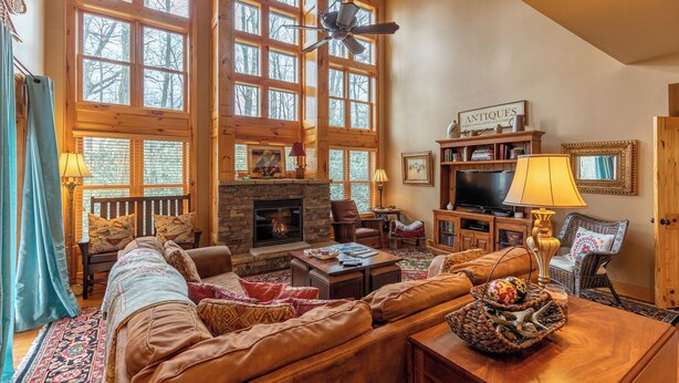 A cozy living room with a fireplace, large windows, a television, and a bookshelf.