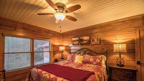 A wooden cabin bedroom with a bed, two nightstands, a ceiling fan, and a window with blinds. - Alpine Vista (Lenoir)