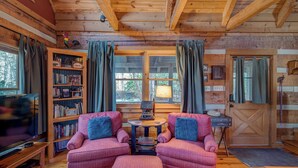 A cozy cabin interior with two upholstered chairs, a wooden bookshelf, and a small table with a lamp. - Scarborough Fair (Banner Elk)