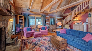 A cozy log cabin interior with wooden beams, a staircase, a fireplace, and a large rug. - Scarborough Fair (Banner Elk)
