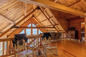 A spacious attic room with wooden beams, a high ceiling, and large windows. There are two rocking chairs and a dining table with chairs. - Beary Relaxing Cabin (Banner Elk)