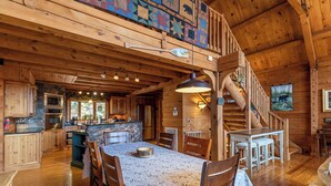 Interior de una cabaña de madera con comedor, cocina y una escalera que sube a un altillo.