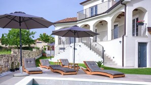 Zona de piscina con sillones de descanso y sombrillas, un muro de piedra y un edificio blanco con balcones y una escalera.