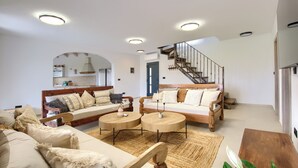 Amplia sala de estar con sofá, dos mesas redondas tipo centro y un banco de madera. Una escalera con barandilla de madera conduce a un nivel superior.