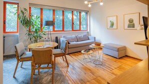 A modern living room with a wooden floor, a round dining table, a grey sofa, and a window with orange frames.