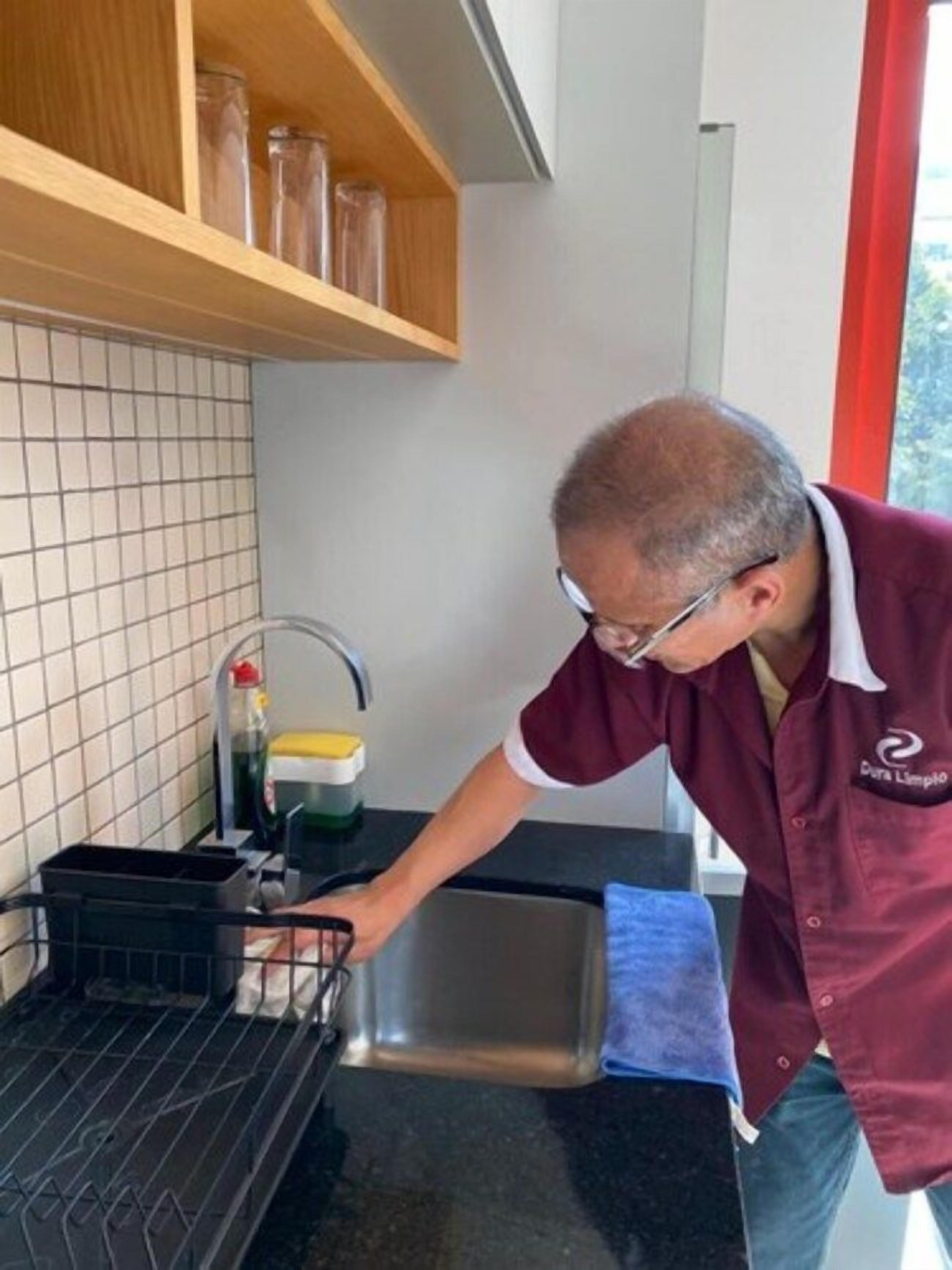 A person in a maroon shirt washing dishes in a kitchen with a tiled backsplash, a wooden shelf, and a window with a red frame.
